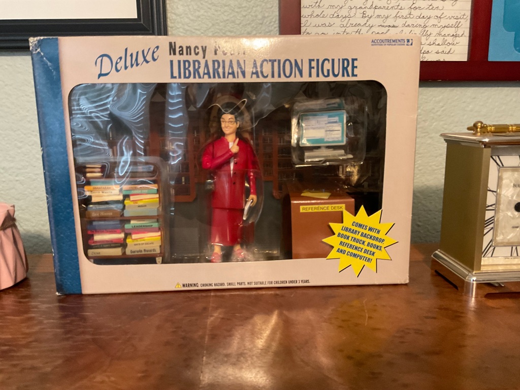 A librarian action figure with accessories. The librarian is wearing a red suit. The accessories include a desk, a cart full of books, a computer, and a library backdrop. 